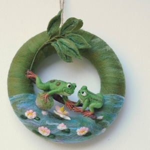 Puede incluir: Una corona de fieltro verde con dos ranas de fieltro verde sentadas en una rama con una hoja de fieltro verde detrás de ellas. La corona está decorada con fieltro azul y blanco para parecer agua y flores de fieltro rosa.