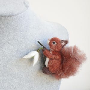 May include: A needle felted red squirrel holding a white snowdrop flower. The squirrel is sitting on a gray surface.