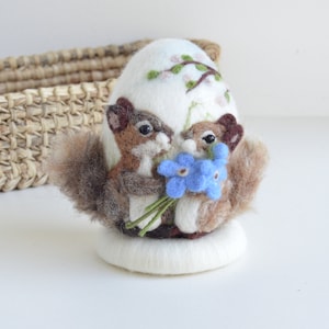May include: Two brown and white felt squirrels holding a bouquet of blue forget-me-not flowers. The squirrels are sitting on a white felt egg with a green branch and pink flowers embroidered on it.