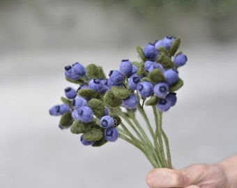 May include: A bouquet of felt blueberries with green leaves and stems. The blueberries are a light purple colour and the leaves are a dark green.
