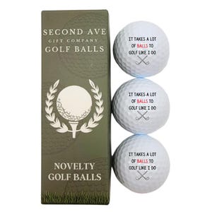 May include: Three white dimpled golf balls and a tall olive green rectangular box. Each ball features the black text IT TAKES A LOT OF BALLS TO GOLF LIKE I DO with the word BALLS in red and a graphic of two crossed golf clubs. The box displays the same text.