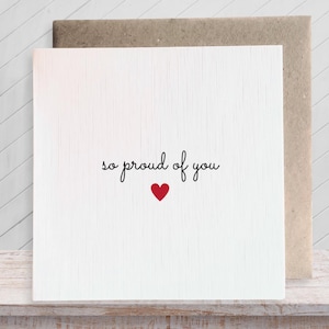 May include: A square white greeting card with a subtle vertical texture. The text "so proud of you" is printed in a black cursive font in the centre, positioned directly above a small, solid red heart icon. The card is paired with a brown envelope.
