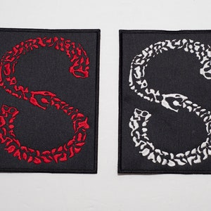 May include: Two black fabric patches with an embroidered white and red snake skeleton in the shape of the letter "S".
