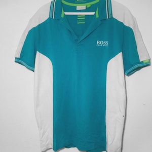 Könnte beinhalten: Türkis-weißes Poloshirt mit dem Logo "BOSS HUGO BOSS" auf der Brust. Das Shirt hat einen Kragen mit grünen und blauen Streifen und weiße Ärmel. Es hängt an einem weißen Kleiderbügel.