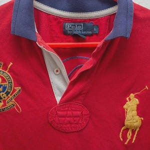 Può includere: Polo rosso con colletto blu navy e dettagli bianchi. Presenta loghi ricamati, tra cui un giocatore di polo dorato e uno stemma con testo. La camicia ha una paramontura abbottonata ed è etichettata "Polo by Ralph Lauren."