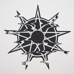 May include: Black and white embroidered patch with a starburst design. The center of the patch is a black circle with a white starburst design inside. The starburst design is surrounded by a white circle with a black starburst design inside.