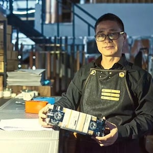 May include: A man wearing a black shirt and a black denim apron with gold accents is holding a box with a black and white design. The box has the text "SINCE" on it.