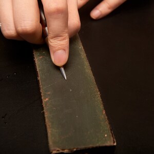 May include: A person's hand using a small, silver tool to sharpen a dark green sharpening stone.