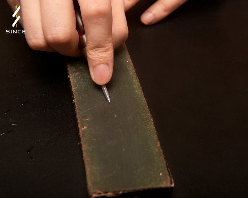 May include: A person's hand using a small, silver tool to sharpen a dark green sharpening stone.