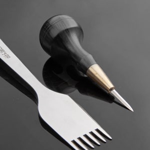 May include: A close-up of two leather working tools. A black handled awl with a brass ferrule and a silver stitching tool with six prongs.