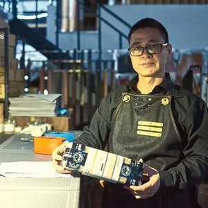 May include: A man wearing a black shirt and a black denim apron with gold accents is holding a box with a black and white design. The box has the text "SINCE" on it.