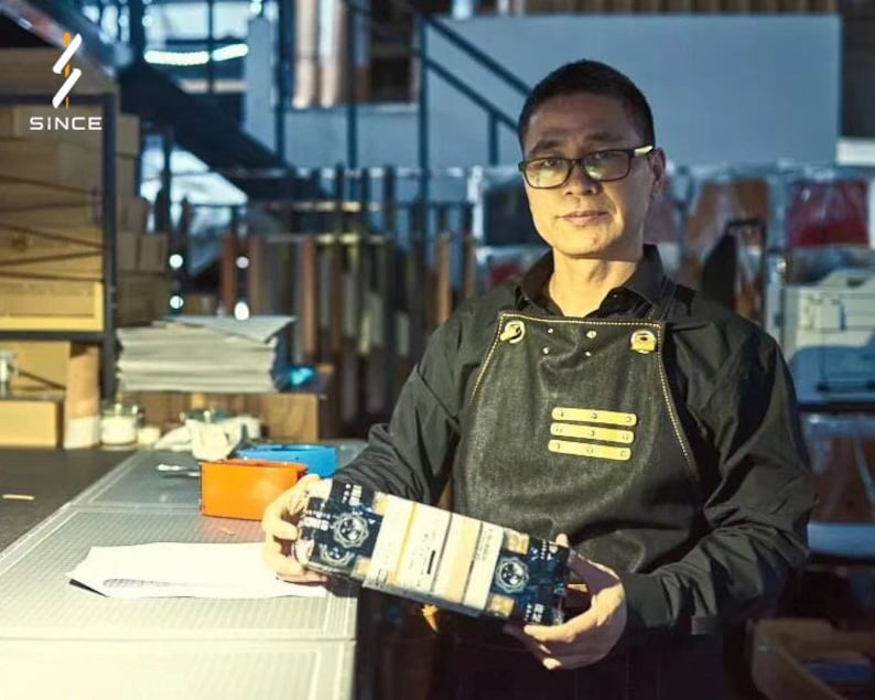 May include: A man wearing a black shirt and a black denim apron with gold accents is holding a box with a black and white design. The box has the text "SINCE" on it.