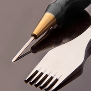 May include: A close-up of a leather crafting tool with a sharp, pointed awl and a metal stitching tool with five prongs. The tools are on a reflective surface.
