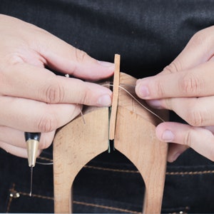 May include: A close-up of a leatherworking tool used for stitching leather. The tool is made of wood and has a groove for holding the leather in place. A needle is being used to thread a piece of leather through the groove.