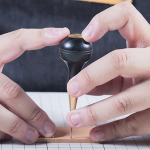 May include: A close-up of a black leather awl with a gold tip being held by a person's hands. The awl is being used to punch a hole in a piece of leather.