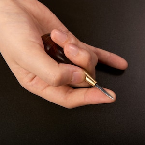 May include: A hand holding a small, wooden-handled leather awl with a sharp, pointed metal tip. The awl is used for piercing holes in leather.