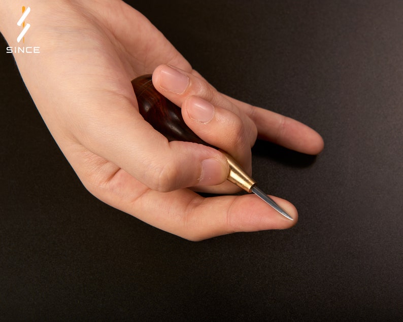 May include: A hand holding a small, wooden-handled leather awl with a sharp, pointed metal tip. The awl is used for piercing holes in leather.