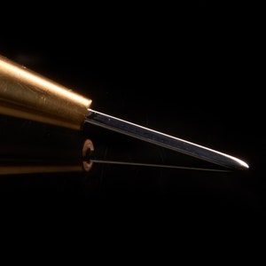 May include: A close-up of a brass awl with a sharp, pointed tip. The awl is a tool used for piercing holes in leather or other materials.