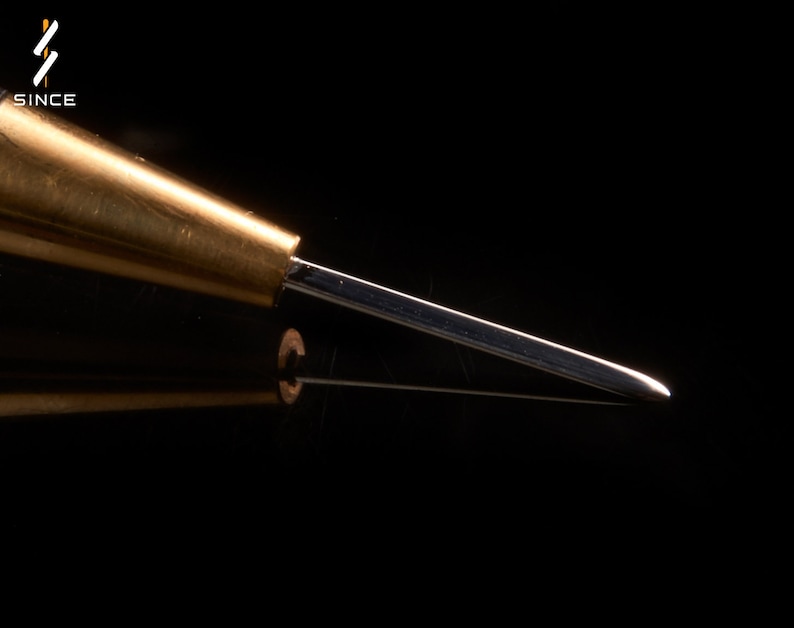 May include: A close-up of a brass awl with a sharp, pointed tip. The awl is a tool used for piercing holes in leather or other materials.