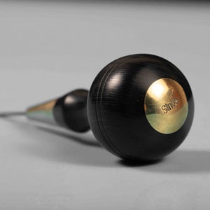 May include: A black wood and brass awl with a round handle. The handle has a brass plate with the word "Since" engraved on it.