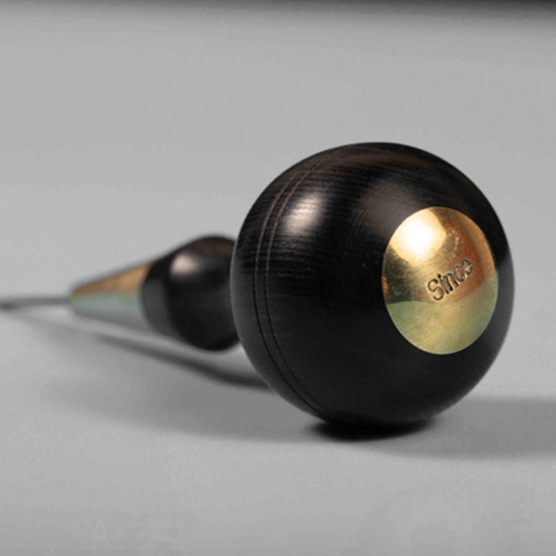 May include: A black wood and brass awl with a round handle. The handle has a brass plate with the word "Since" engraved on it.