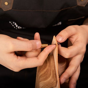 May include: Close-up of a person's hands using a leather stitching tool to sew two pieces of brown leather together. The leather is being held in place with a white thread.