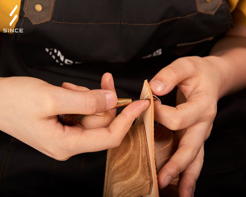 May include: Close-up of a person's hands using a leather stitching tool to sew two pieces of brown leather together. The leather is being held in place with a white thread.