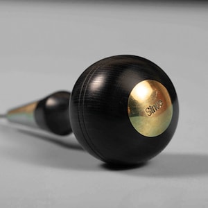May include: A black wood and brass awl with a round handle. The handle has a brass disc with the word "Since" engraved on it.