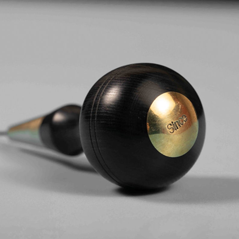 May include: A black wood and brass awl with a round handle. The handle has a brass disc with the word "Since" engraved on it.