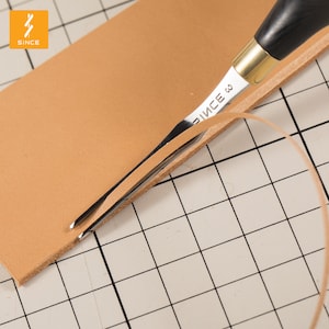 Può includere: Primo piano di uno strumento per la lavorazione del cuoio con manico in legno e lama in metallo. Lo strumento viene utilizzato per tagliare un pezzo di cuoio marrone. Il cuoio è su una superficie a griglia.