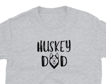 May include: A light gray t-shirt with the text "HUSKEY DAD" in a black, handwritten-style font. The word "DAD" has a husky dog face in the middle. The shirt is made of a soft material.