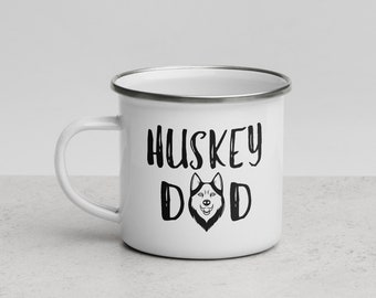 Puede incluir: Taza de esmalte blanco con borde y asa plateados. La taza presenta el texto "HUSKEY DAD" en negro, con una ilustración estilizada de un perro husky en el centro. La taza está diseñada para bebidas calientes.