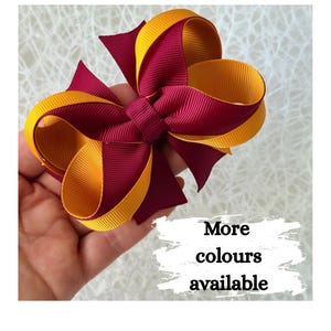 May include: A handmade hair bow in burgundy and gold. The bow is held in a hand and has the text "More colours available". The bow is made of layered ribbons.