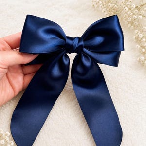 May include: A dark navy blue hair bow crafted from smooth, lustrous satin ribbon. The accessory features a central knot with two wide, structured loops at the top and two long, flowing tails extending downward with angled, finished edges.