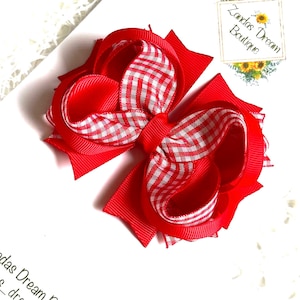 May include: A bright red hair bow with a white and red gingham centre. The bow is made of layered ribbons and features the text "Zandas Dream Boutique" on a small card.