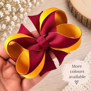 May include: A hair bow made of layered maroon and yellow grosgrain ribbon with a central knot. The bow features multiple loops and pointed ends. Text on the image reads: More colours available with a small heart symbol below the text.