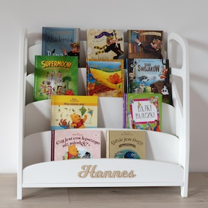May include: A white wooden bookshelf filled with colorful children's books. The bookshelf has a curved design with multiple shelves. The name "Hannes" is displayed on the front of the bookshelf.