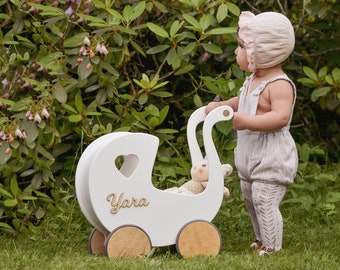 May include: A white wooden doll's pram with a heart-shaped cutout and the name "Yara" written on the side. A young child in a pink bonnet and white overalls is pushing the pram.