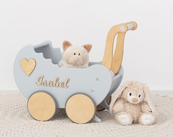May include: A light blue wooden doll's pram with a wooden handle and wheels. The pram has a heart-shaped cutout and the name "Isabel" written on the side. There are two plush toys, a light brown bunny and a white and brown pig, sitting in and next to the pram.