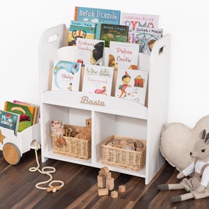 May include: White wooden bookshelf with two shelves and a top section for books. The bookshelf has a cutout handle on the side and a nameplate that reads "Basia". The shelves are filled with children's books and toys, including a toy wagon, stuffed animals, and wooden blocks.