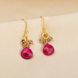 Puede incluir: Un par de pendientes dorados con un grupo de pequeñas cuentas grises sobre una gran gema magenta en forma de lágrima. Los pendientes se muestran sobre una tela de color crema.