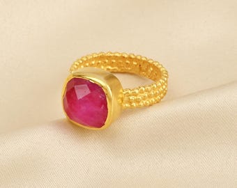 May include: A gold-colored ring with a square, faceted, magenta gemstone. The ring band has a textured, beaded design. The ring is set against a cream-colored, textured background.