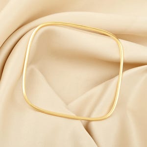 May include: A gold-colored, square bangle bracelet. The bracelet is a simple, minimalist design with a smooth, polished finish. The jewelry is displayed on a cream-colored, textured fabric.