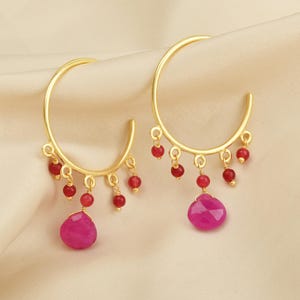 Pode incluir: Um par de brincos de argola dourados. Cada brinco apresenta pequenas contas vermelhas penduradas na parte superior e uma grande pedra rosa facetada em forma de gota na parte inferior. Os brincos são exibidos em uma superfície creme.