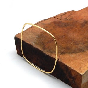 Op de afbeelding: Een goudkleurige, vierkant gevormde armband. De armband heeft een gehamerde textuur en wordt gepresenteerd op een bruin houten oppervlak. Het ontwerp van de armband is minimalistisch en modern.