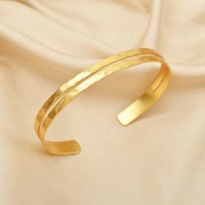 Peut inclure: Un bracelet manchette doré avec une texture martelée. Le design ouvert présente deux bandes parallèles qui se courbent pour former le bracelet. Le bijou est présenté sur une surface en satin crème.