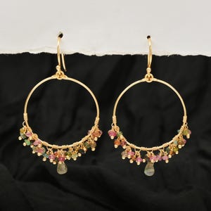 Puede incluir: Un par de pendientes de aro dorados con cuentas de piedras preciosas de colores. Los pendientes presentan un dise&ntilde;o de aro circular grande con peque&ntilde;as cuentas multicolores y una sola cuenta en forma de l&aacute;grima colgando de la parte inferior.