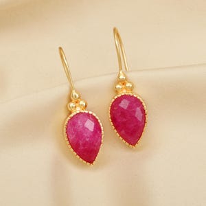 Op de afbeelding: Een paar gouden oorhangers met roze druppelvormige stenen. De stenen zijn gefacetteerd en hebben een gouden rand.