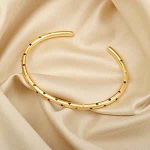 Könnte beinhalten: Ein goldenes Armreif-Armband mit einem dünnen Band und einer Reihe von bunten Edelsteinen am Rand.