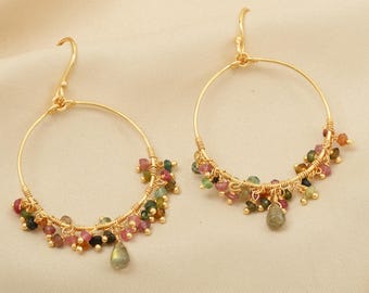 May include: A pair of gold-colored hoop earrings. Each earring features a gold wire design with clusters of small, colorful beads in shades of pink, green, and brown. A teardrop-shaped bead hangs from the bottom of each earring.